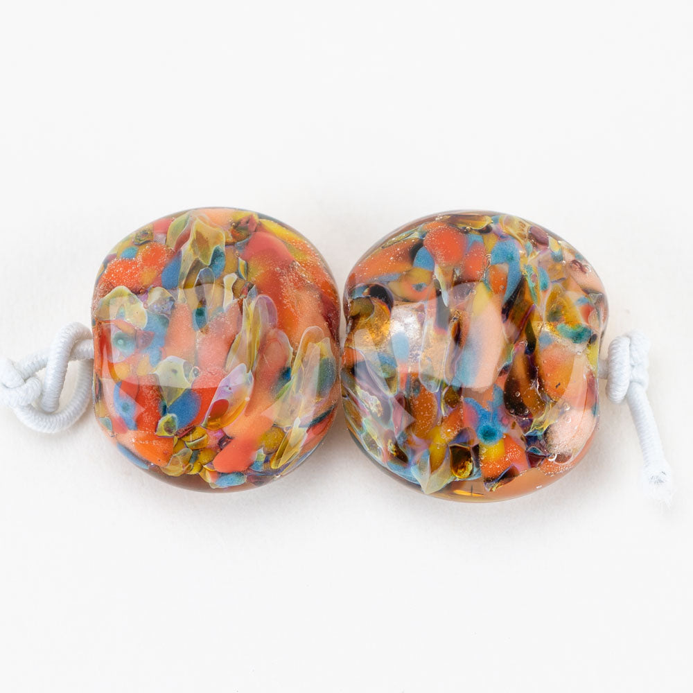 Two glass beads from Unicorne Beads. Each bead has a mix of colors including orange, blue, red, and brown. Each bead has a hole through it to be used for jewelry projects.