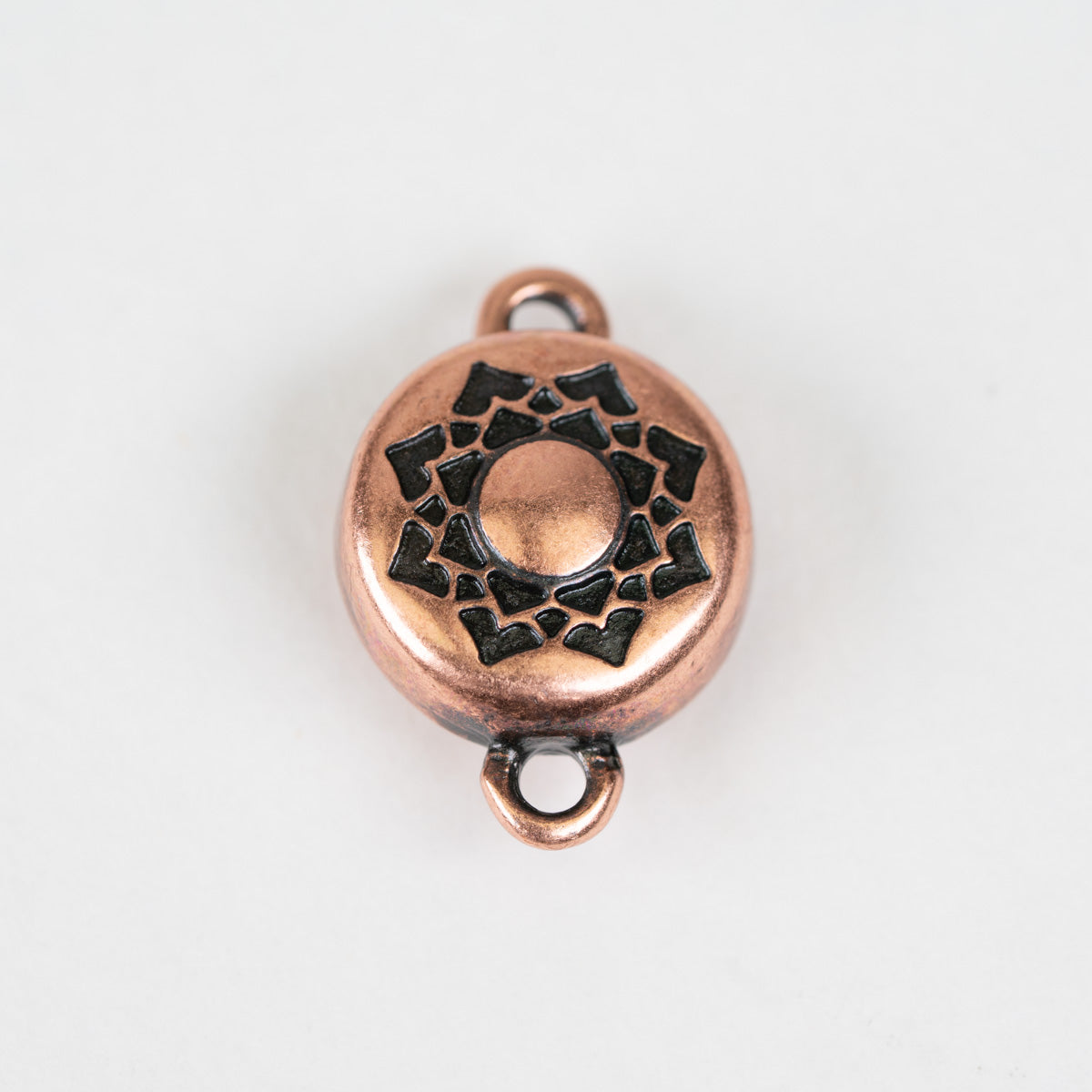 A round copper-colored metal charm with a black engraved geometric sunburst design in the center, featuring two small loops at the top and bottom, shown on a plain white background.