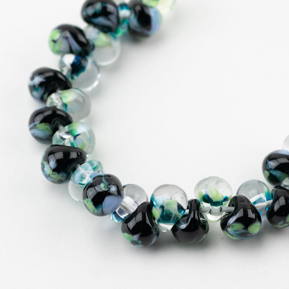 A strand of Fairbanks Teardrop beads crafted by Unicorne Beads. These Beads are shaped like teardrops and are black  with green accents. Each bead has a hole in the middle to be used for jewelry craft projects.