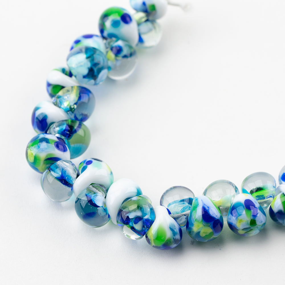 A strand of Zurich Teardrop beads crafted by Unicorne Beads. These Beads are shaped like teardrops and are white, blue, and green. Each bead has a hole in the middle to be used for jewelry craft projects.