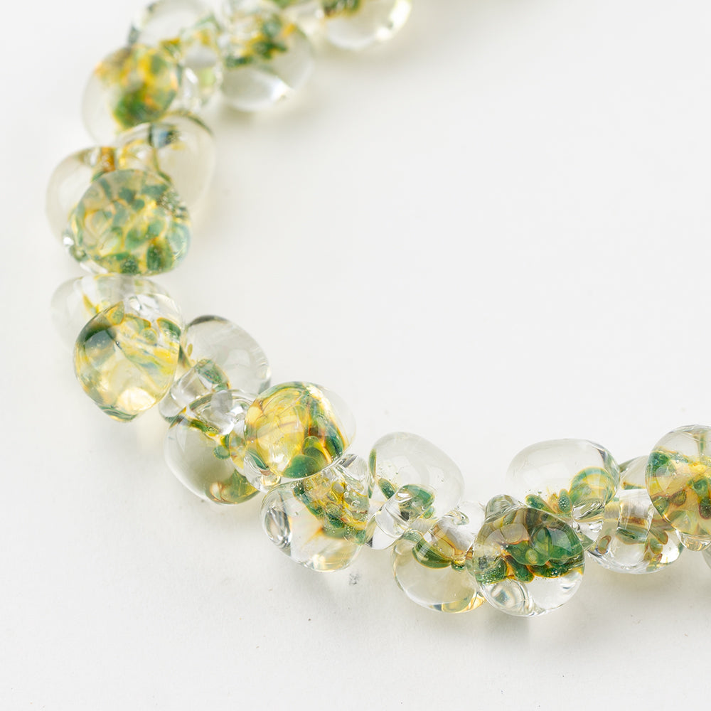 A strand of Luminescence Teardrop glass beads crafted by Unicorne Beads. These glass beads are shaped like teardrops and are green and gold. Each glass bead has a hole in the middle to be used for jewelry craft projects.