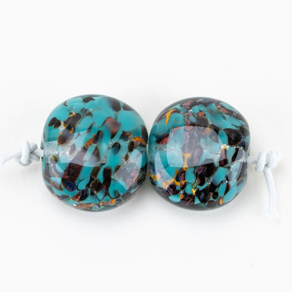 Two glass beads from Unicorne Beads. Each bead features a teal and copper color scheme and have holes on either side to be used for jewelry projects.