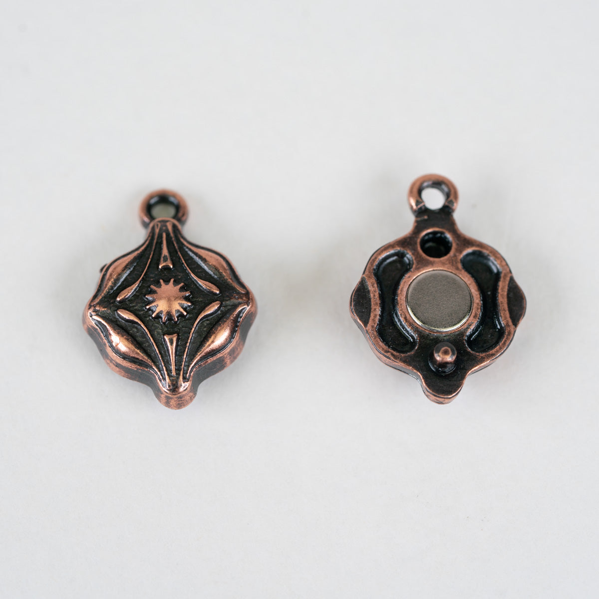 Two small decorative metal charms or pendant components shown on a plain light background, one facing forward with an engraved starburst motif and the other showing the reverse side with a circular opening.
