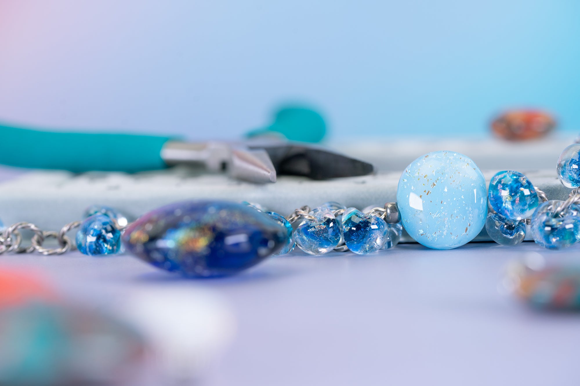 A close-up of handmade beaded jewelry featuring light blue, deep blue, and iridescent blue glass beads with silver-toned metal links, set against a soft pastel blue and lavender background with blurred craft tools in the distance.