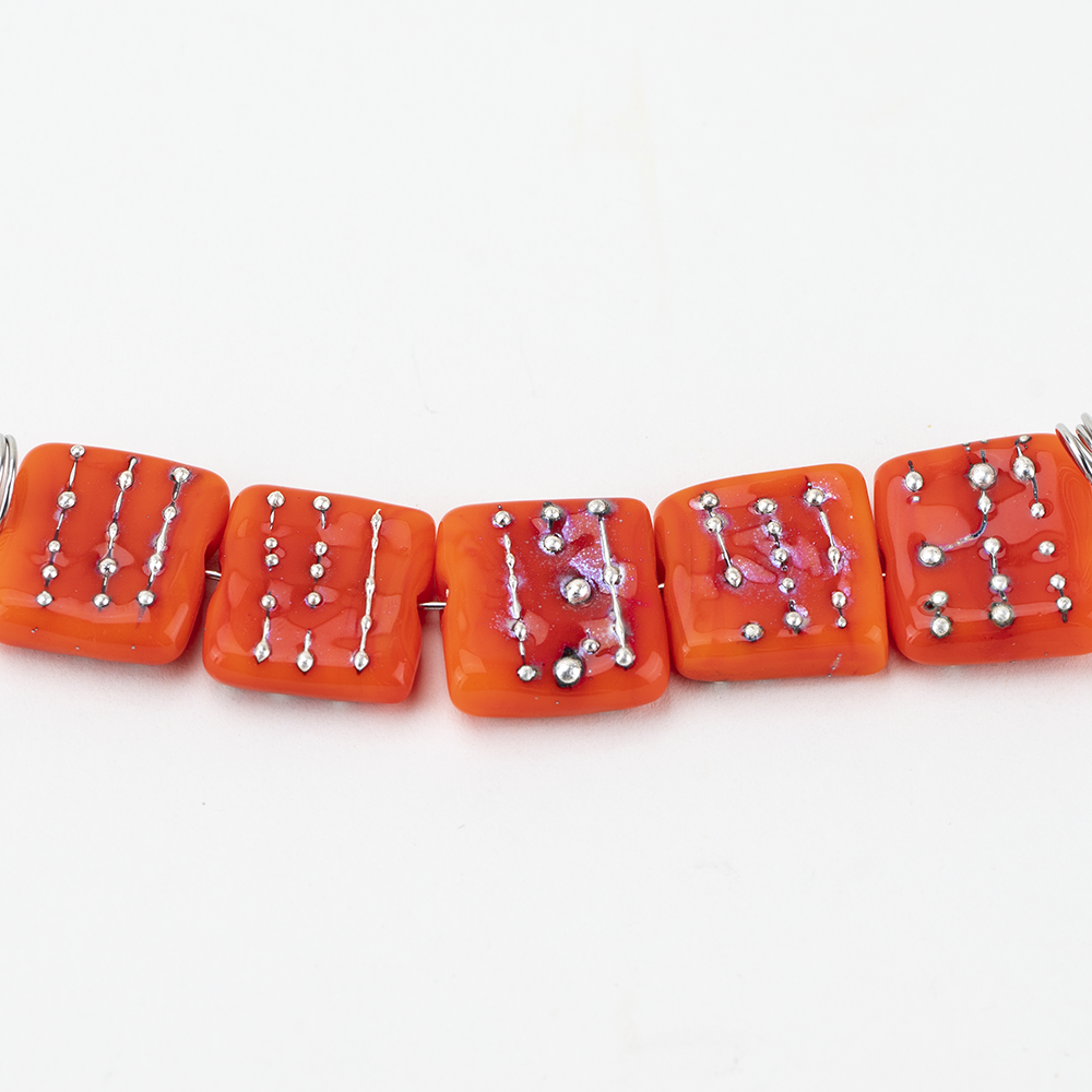 Five tile glass beads blaze orange in color. Each bead is sqaure in shape and features silver globs embedded in the glass. A string runs through the glass beads to be used for jewelry.