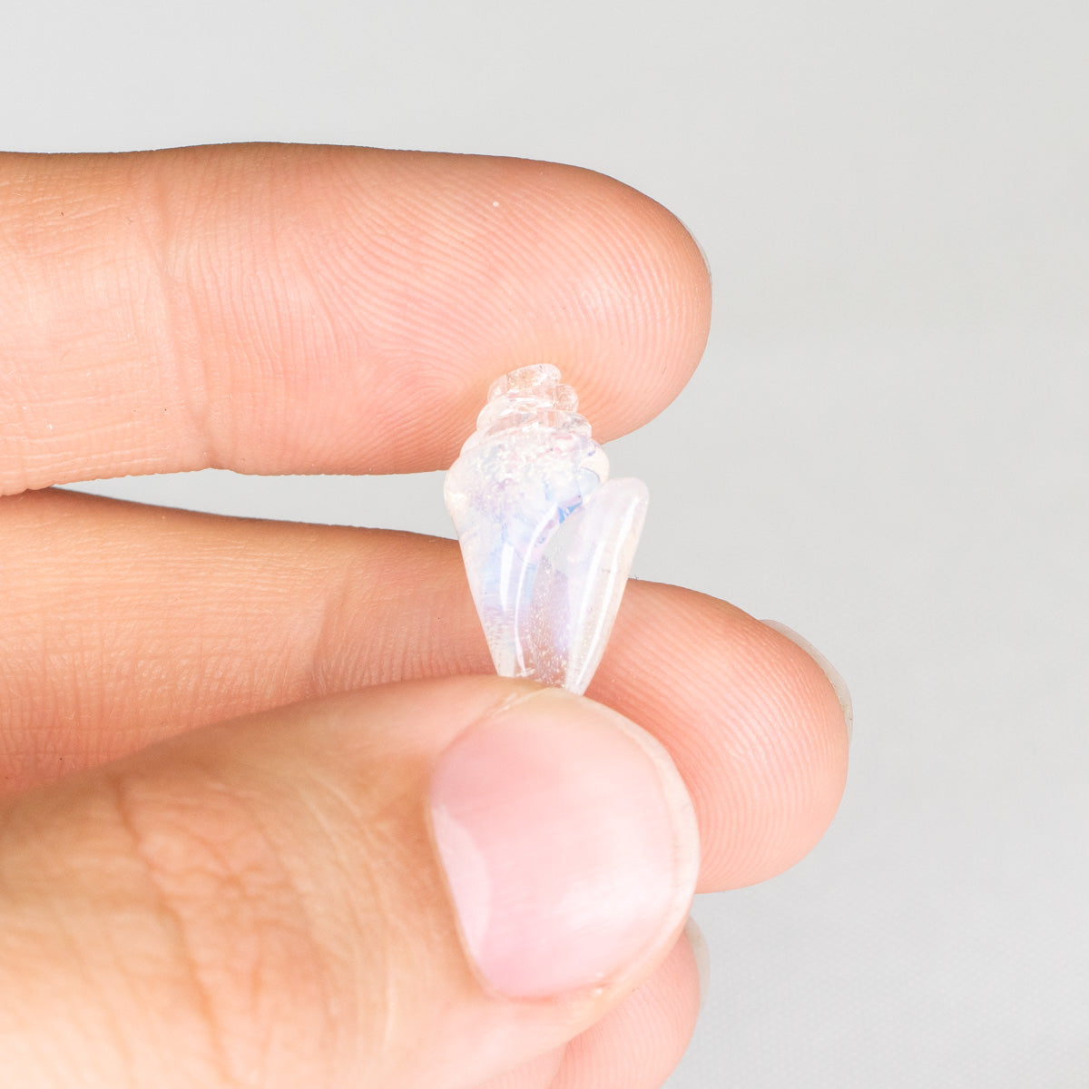 Hand holding a small, clear seashell bead against a neutral background