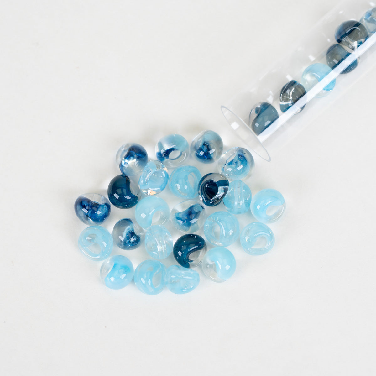 Light blue and dark blue marbled glass beads spilled from a clear plastic tube on a white background.