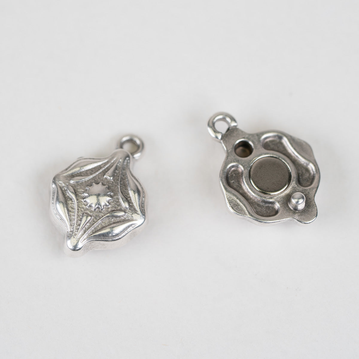 Two silver-toned metal jewelry findings or charms shown on a plain light background, one with an ornate open star-like design and a clear stone, and the other showing the back with a circular magnetic area and small loops.