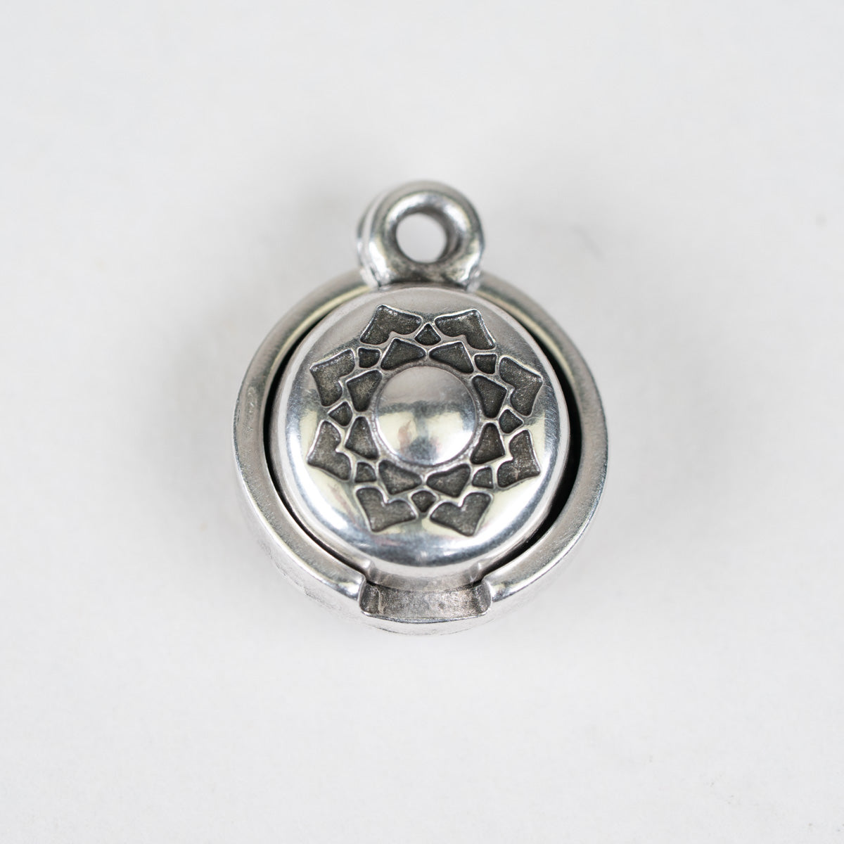 A round silver-toned metal charm or pendant with a textured geometric starburst design on the front and a small loop at the top, shown on a plain light background.