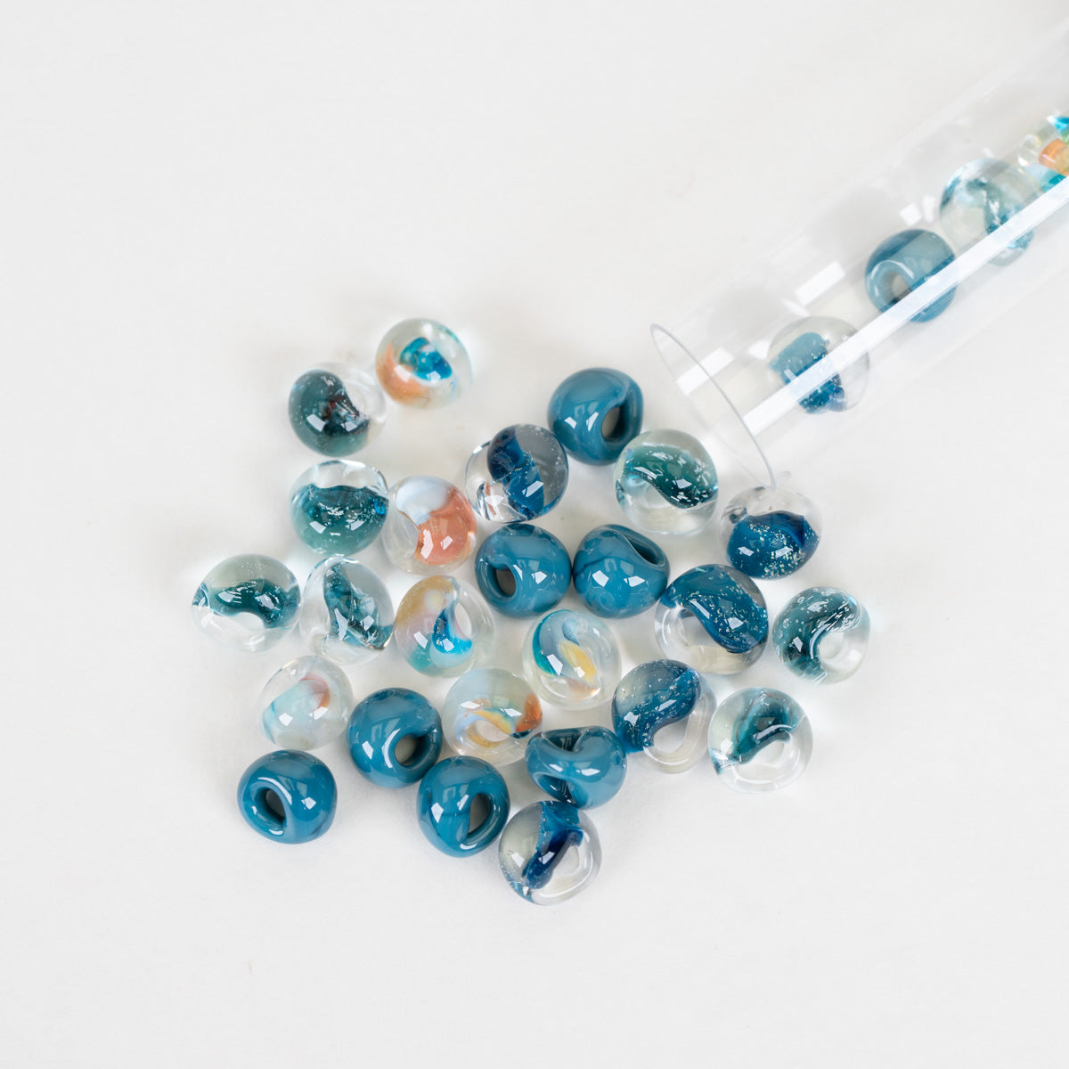 Blue and clear glass beads with swirls of teal, navy, and orange, spilling from a clear glass tube onto a white surface.