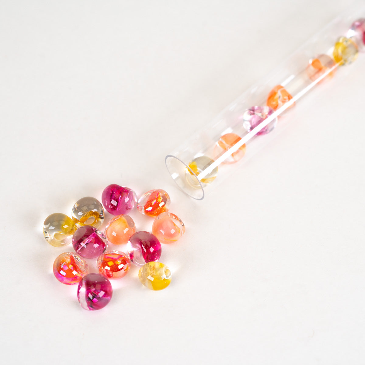 A clear glass tube filled with colorful faceted beads, with several beads spilled out on a plain white surface. The beads are translucent and come in shades of pink, orange, yellow, and clear.