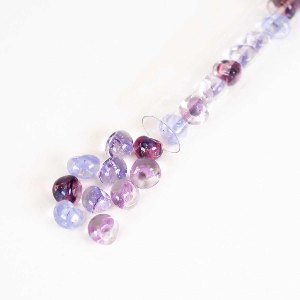 Faceted glass beads in shades of purple, lavender, and clear, with some spilled from a clear plastic tube on a white background.
