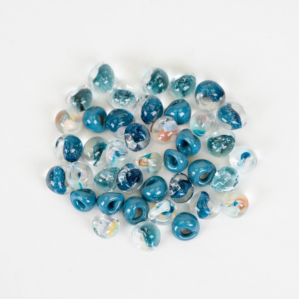 A pile of small, teardrop-shaped glass beads in shades of blue, clear, and white, with some containing tiny orange inclusions, set against a plain white background.