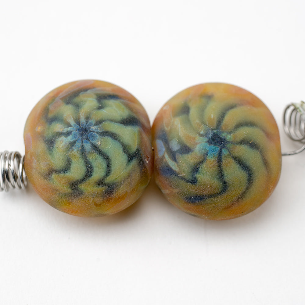 Two glass beads featuring an orange and green spiral pattern. Each bead is matte in finish and has a hollow center to be used for jewelry making.