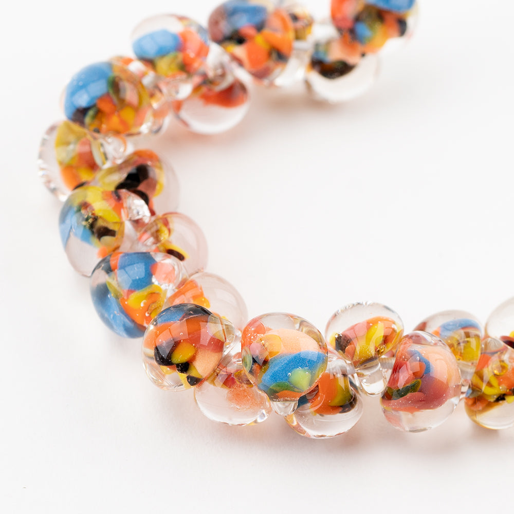 A strand of Bird of Paradise Teardrop glass beads crafted by Unicorne Beads. These glass beads are shaped like teardrops and are orange, blue, clear, and black. Each glass bead has a hole in the middle to be used for jewelry craft projects.