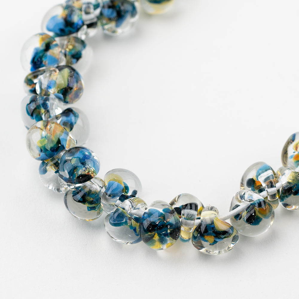 A strand of Coral Flourish glass beads crafted by Unicorne Beads. These glass beads are shaped like teardrops and are Blue and black, with yellow accents. Each glass bead has a hole in the middle to be used for jewelry craft projects.