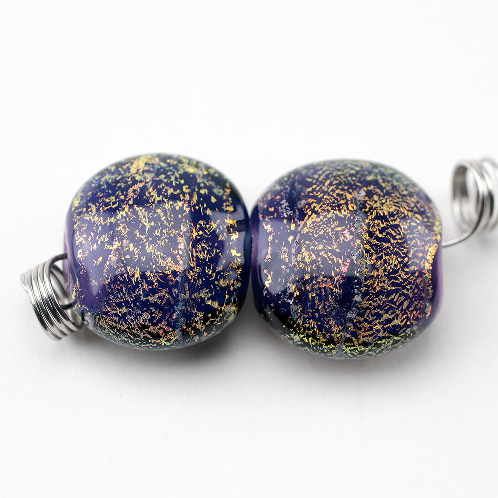 Two premium jewelry beads placed next to each other. Each bead has a dark purple color with dichroic coloring. Each bead features a hollow center where it can be strung on jewelry.