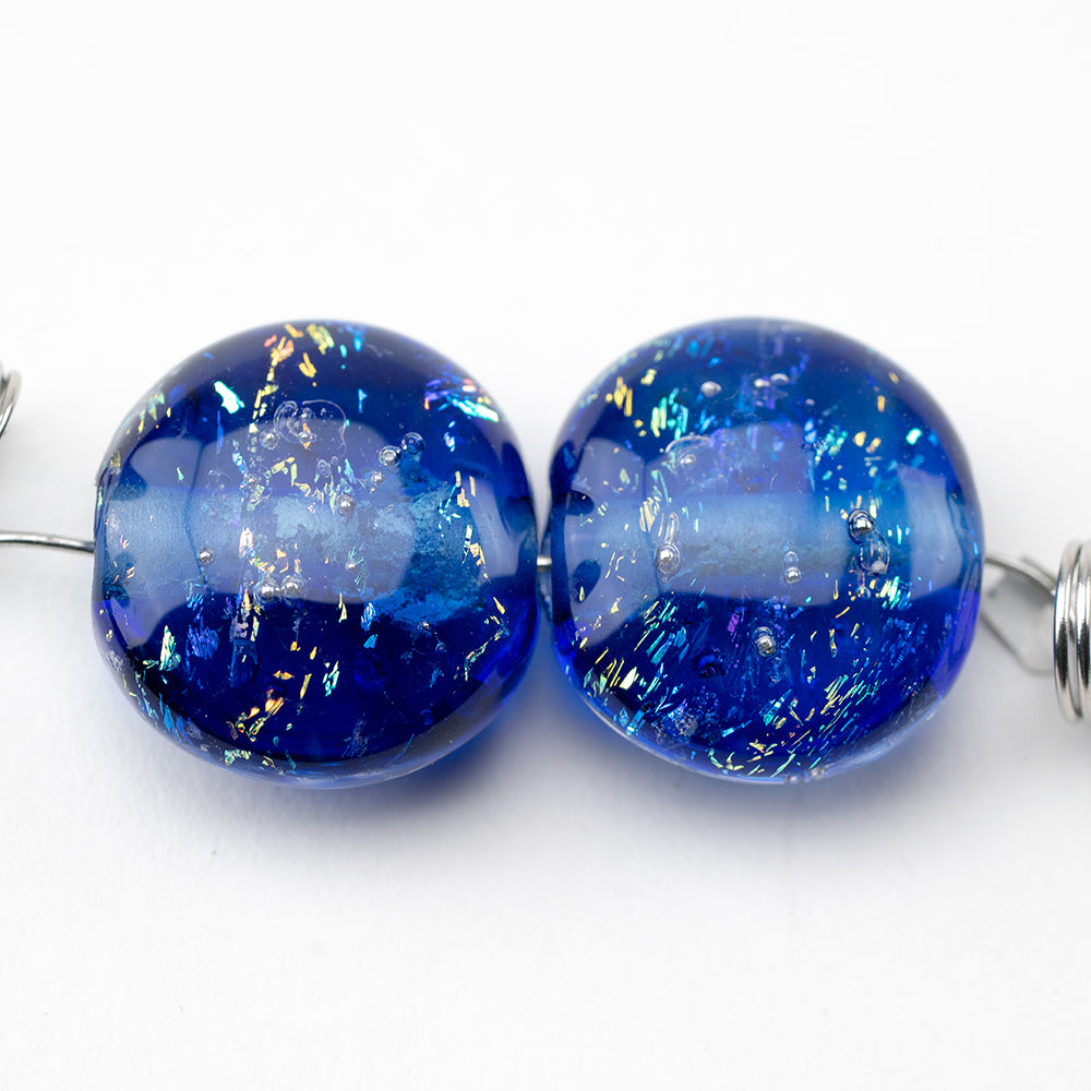Two glass beads dark blue in color with features of dichroic foiling. Each bead has a hollow center with a string going through them.