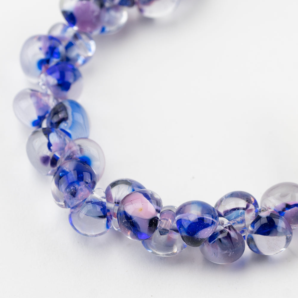A strand of Orsi Teardrop glass beads crafted by Unicorne Beads. These glass beads are shaped like teardrops and are blue, purple, and pink. Each glass bead has a hole in the middle to be used for jewelry craft projects.