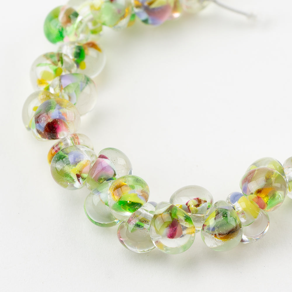 A strand of King Cake Teardrop glass beads crafted by Unicorne Beads. These glass beads are shaped like teardrops and are green and yellow, with multi-color accents. Each glass bead has a hole in the middle to be used for jewelry craft projects.