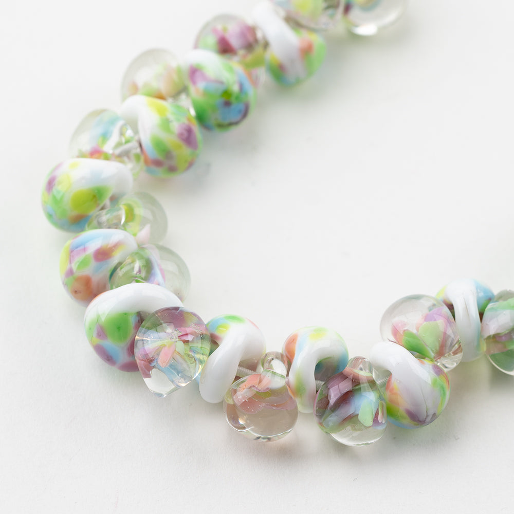 A strand of Peony Teardrop beads crafted by Unicorne Beads. These Beads are shaped like teardrops and are white, clear, green, pink, and blue. Each bead has a hole in the middle to be used for jewelry craft projects.