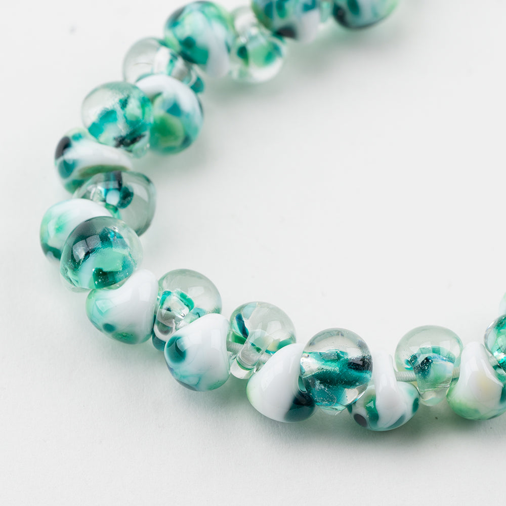 A strand of Helsinki Teardrop beads crafted by Unicorne Beads. These beads are shaped like teardrops and are white, teal, and clear glass. Each bead has a hole in the middle to be used for jewelry craft projects.