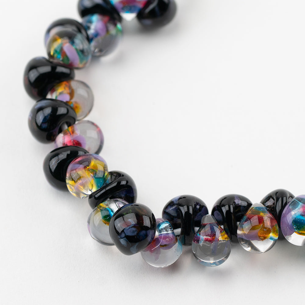 A strand of Minsk Teardrop beads crafted by Unicorne Beads. These Beads are shaped like teardrops and are black and clear with rainbow accent swirls. Each bead has a hole in the middle to be used for jewelry craft projects.