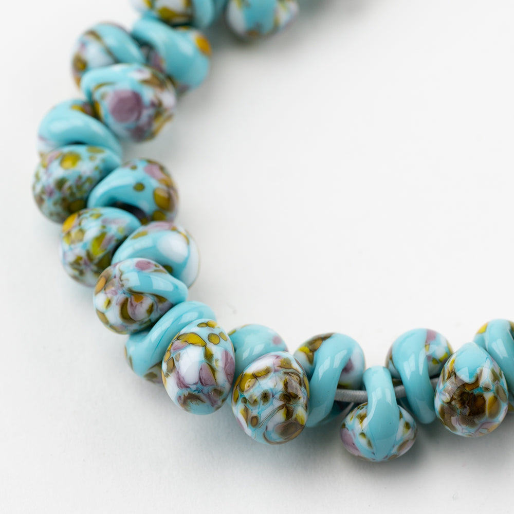 A strand of Antigua Teardrop beads crafted by Unicorne Beads. These Beads are shaped like teardrops and are teal and brown, with a hint of pink. Each bead has a hole in the middle to be used for jewelry craft projects.