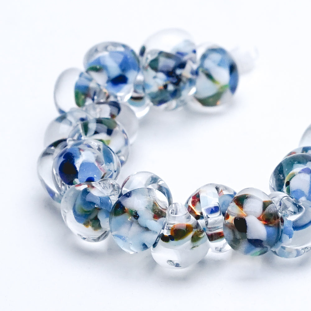 A strand of Blue Poppy glass beads crafted by Unicorne Beads. These glass beads are shaped like teardrops and are blue, purple, with multi-color accents. Each glass bead has a hole in the middle to be used for jewelry craft projects.