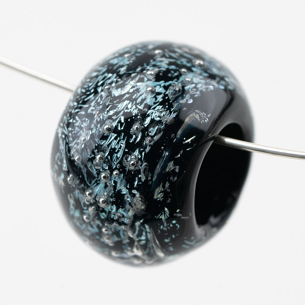 Donut-shaped glass bead black in color with dichroic foil and silver globs. The large bead has a center hole allowing it to be strung on jewelry pieces.