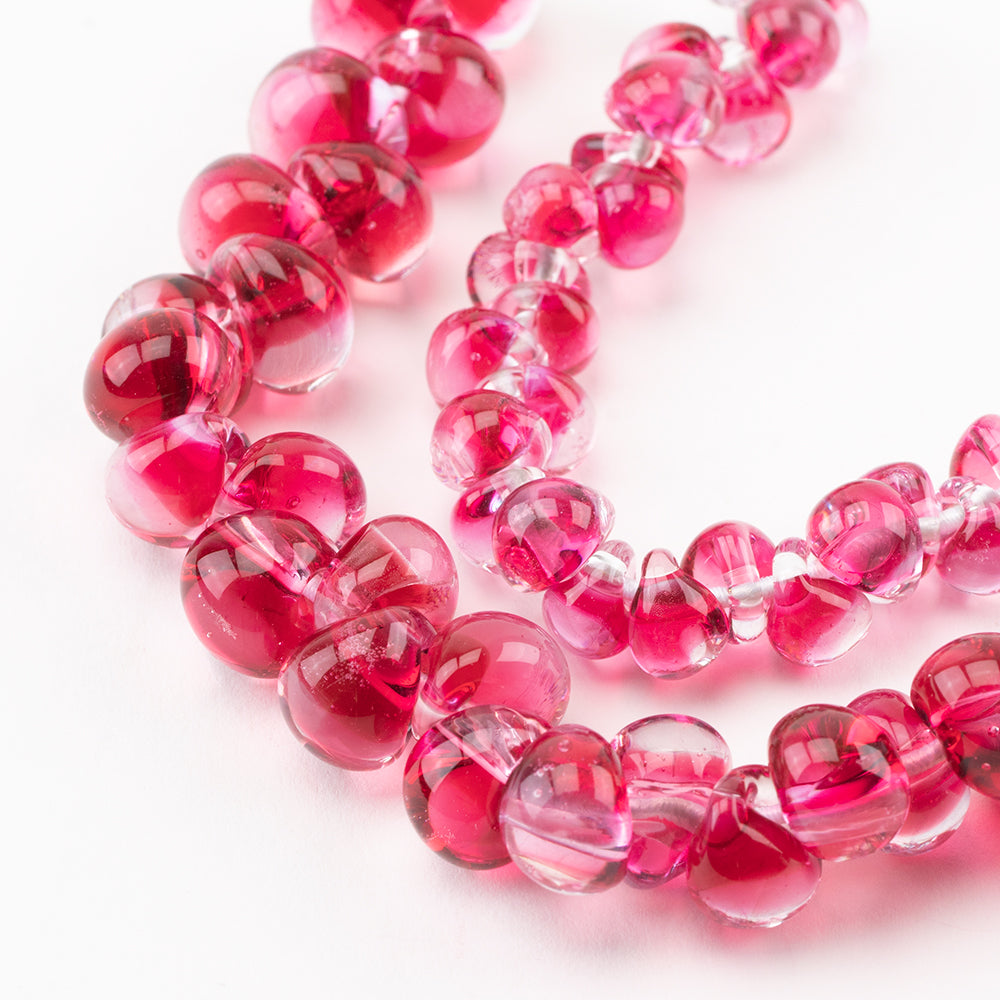 Two seperate strands of glass beads from Unicorne Beads. Both strands are the same hot pink color, mixed with clear glass. The left strand is bigger glass beads and the right strand is mini glass beads. Glass beads are used for jewelry projects.